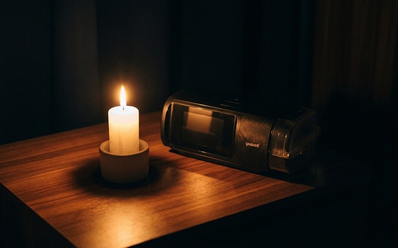 A Yuwell CPAP machine glowing softly on a wooden nightstand beside a lit candle during a nighttime brownout.