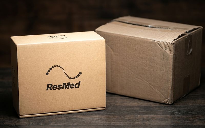 A plain retail ResMed box standing next to a battered, unbranded plain shipping carton of similar size on a dark wooden surface.