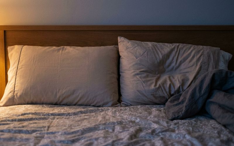 Two pillows on a neatly made bed in a dim bedroom lit by a bedside lamp.
