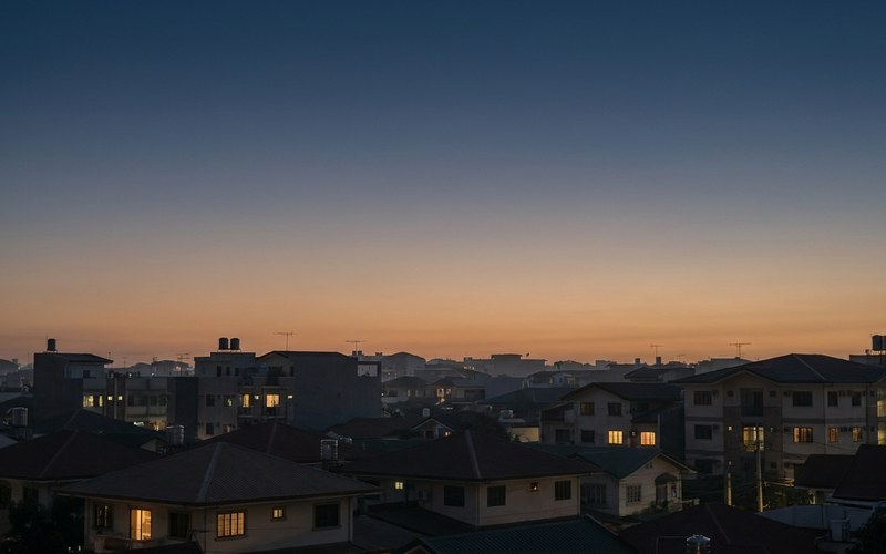A dusk skyline of ordinary houses and low apartment blocks with windows lit warm across many separate homes.